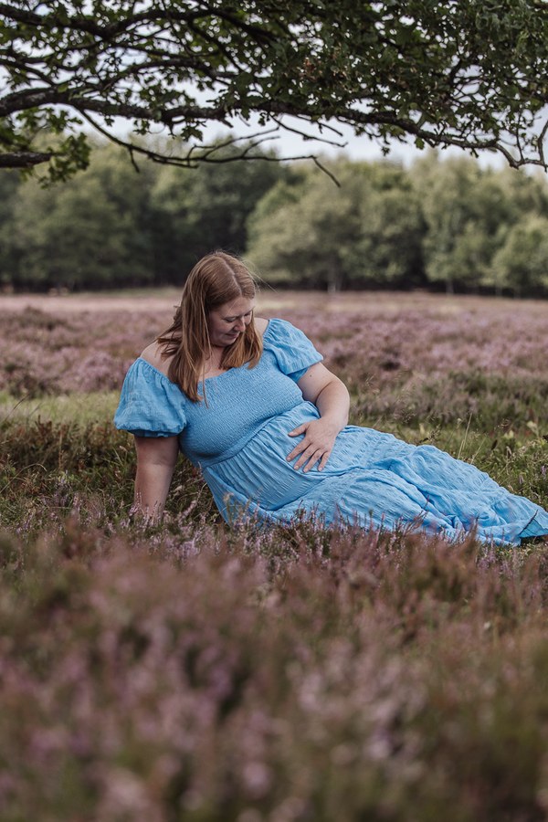 Gravid kvinde i blå kjole ligger i den blomstrende lyng under et træ